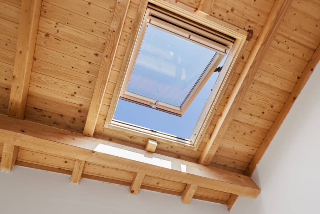 Plafond mansardé en bois clair avec poutres apparentes et grande lucarne de toit ouverte sur un ciel bleu lumineux.