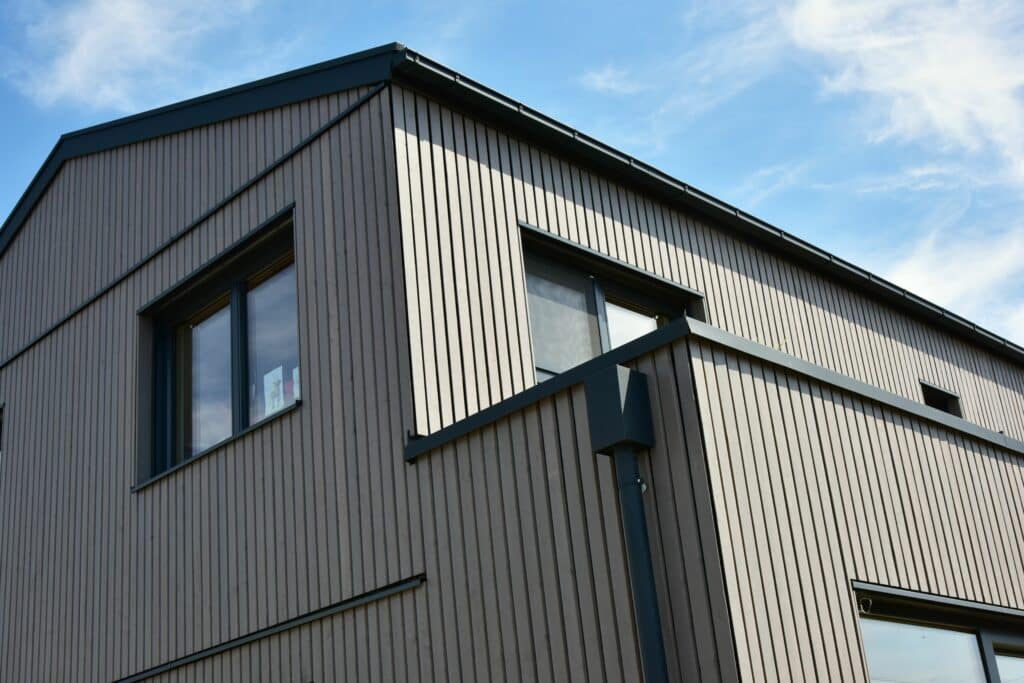 Façade moderne d'un bâtiment en bois gris à lamelles verticales, fenêtres sombres et toiture foncée sous ciel bleu.