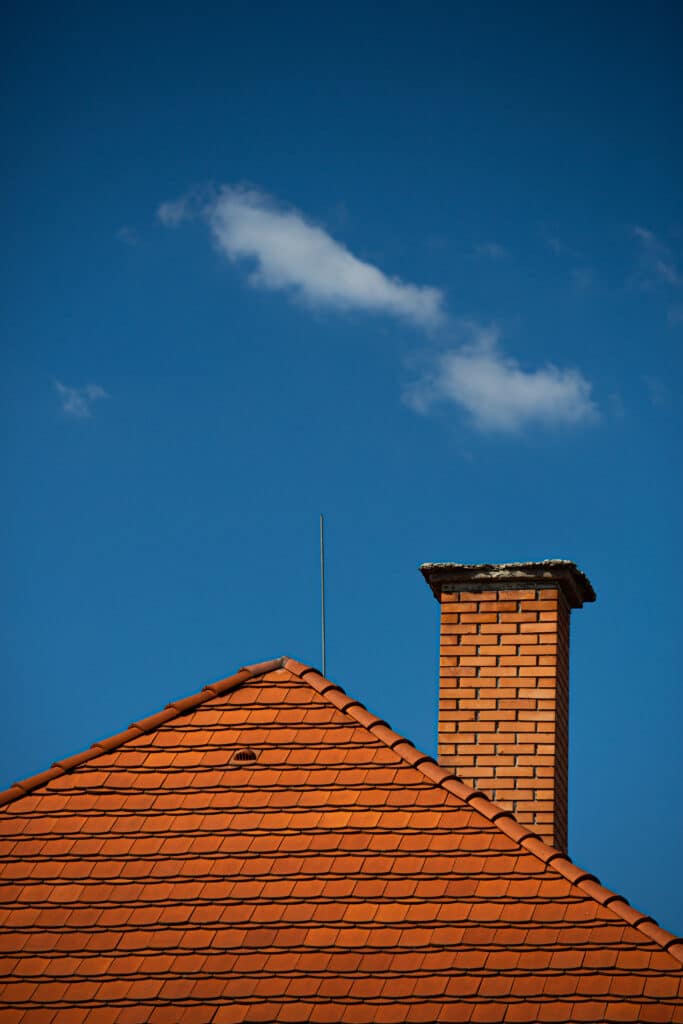 Toit en tuiles orange, cheminée en brique et paratonnerre sous un ciel bleu clair avec quelques nuages blancs.